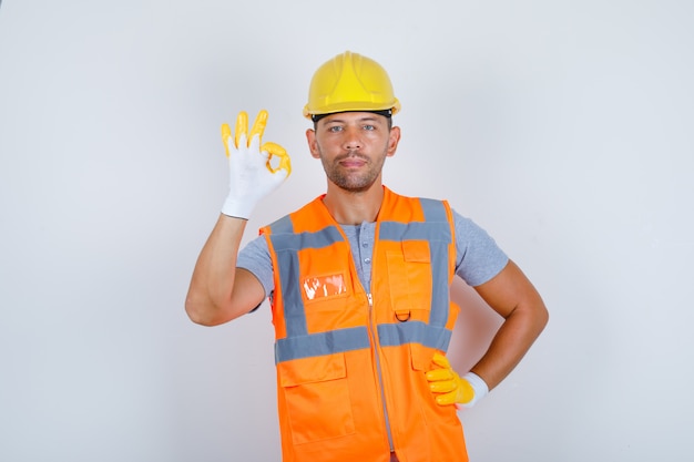 Male builder doing ok gesture with hand on waist in uniform, helmet, gloves, front view.