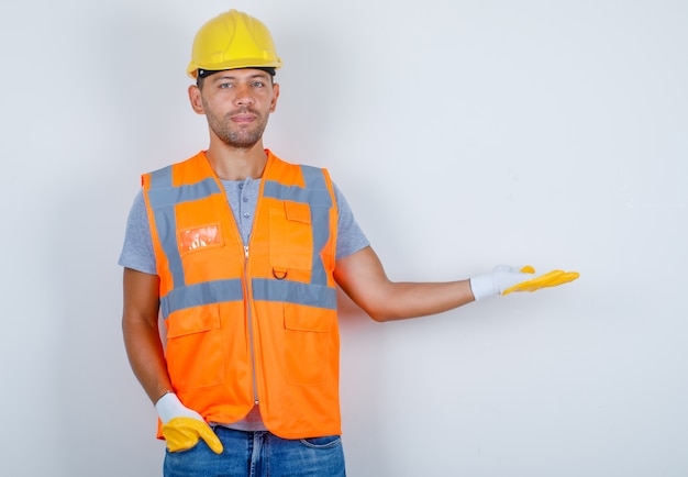 Male builder gesturing as welcoming with hand in pocket in uniform, jeans, helmet, gloves, front view.
