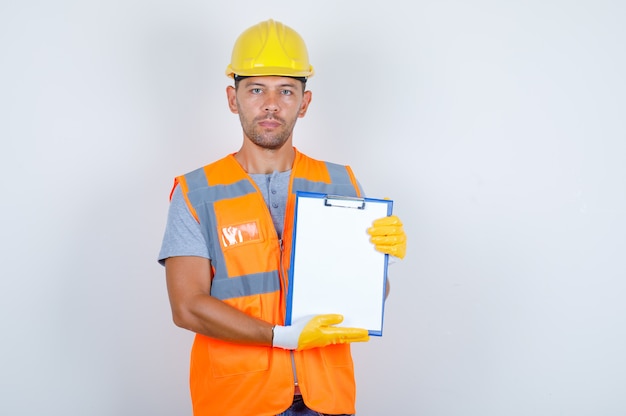 Male builder holding clipboard and looking at camera in uniform, helmet, gloves front view.