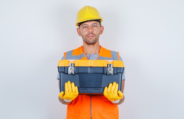 Строитель-мужчина держит пластиковый ящик для инструментов в форме, шлеме, перчатках, вид спереди.