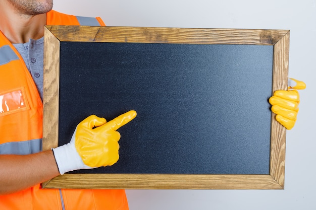 Male builder in uniform, gloves showing something on blackboard, front view.