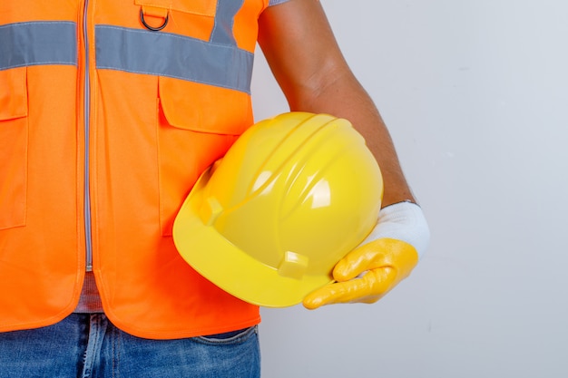 Male builder in uniform, jeans, gloves holding helmet in his hand, front view.