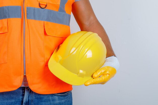 Male builder in uniform, jeans, gloves holding helmet in his hand, front view.