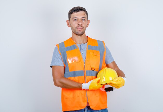 Male builder in uniform, jeans, gloves holding helmet in his hands, front view.