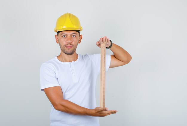 Costruttore maschio in maglietta bianca, casco di sicurezza che tiene il righello di legno, vista frontale.