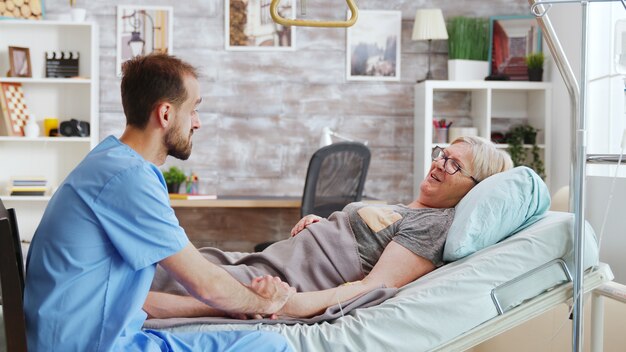 Male caregiver talking with a sick old lady lying in hospital bed, he takes the hand of the woman