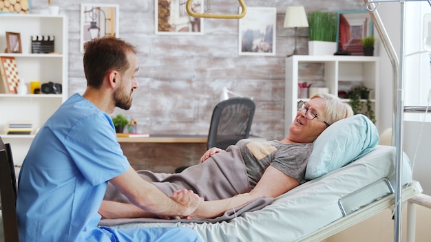 Male caregiver talking with a sick old lady lying in hospital bed, he takes the hand of the woman