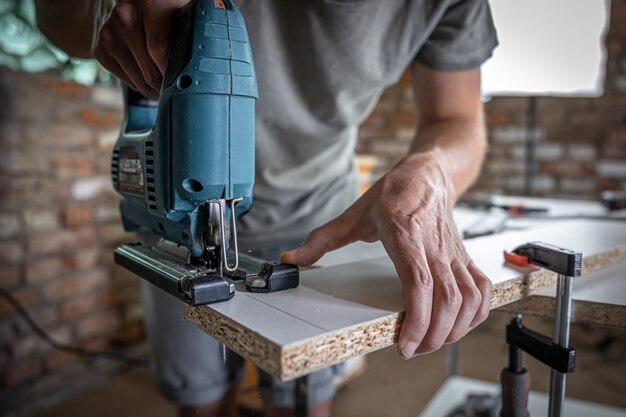 A male carpenter cuts a wood with an electric jigsaw, working with a tree.