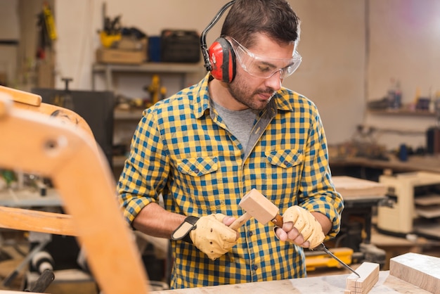 Male carpenter holding a hammer in his hand hitting a chisel on wooden block