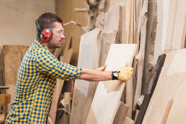 Мужской плотник в защитных очках держит деревянную доску в мастерской