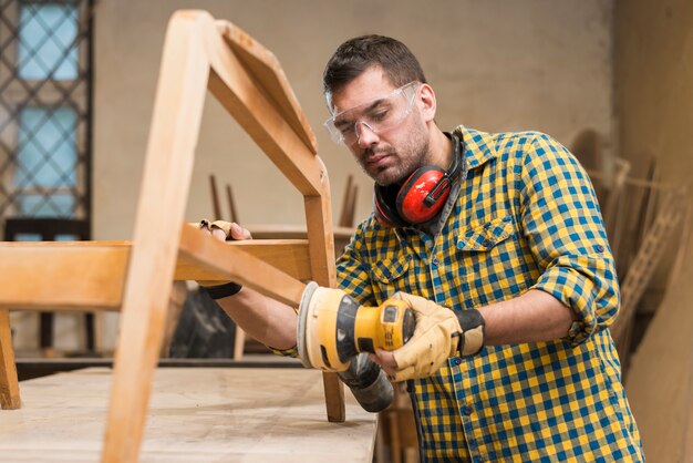 Мужской плотник носить защитные очки с помощью шлифовальной машины на мебель в мастерской