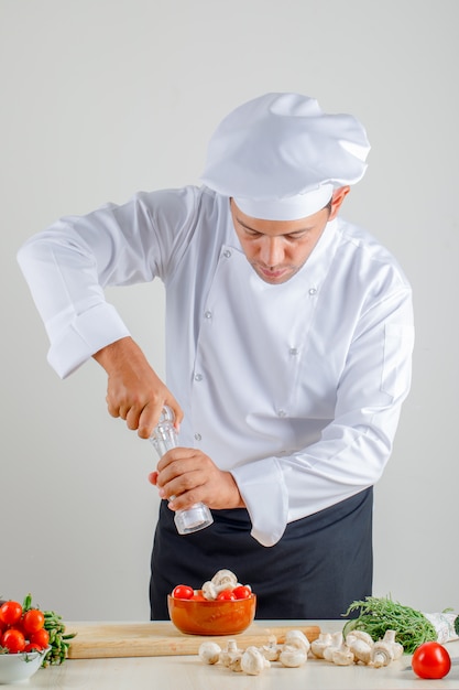 Cuoco unico maschio che aggiunge sale al cibo in uniforme, cappello e grembiule in cucina
