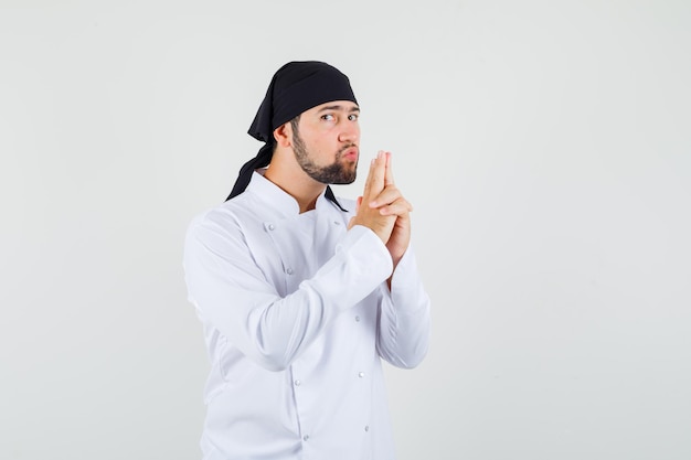 Male chef blowing on gun made by his hands in white uniform and looking confident. front view.