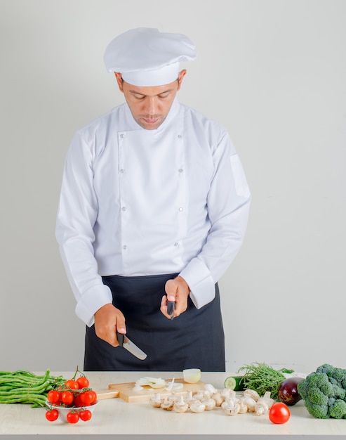 Male chef choosing knife for chopping onion in kitchen in uniform, hat and apron