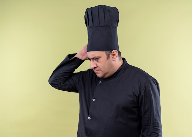 無料写真 男性シェフの料理人は黒い制服を着て、混乱して脇を見て帽子を調理し、緑の背景の上に立っている何かを忘れました