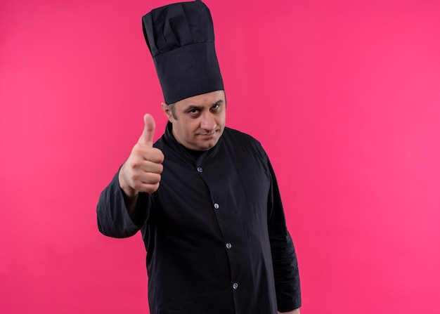 無料写真 男性シェフの料理人は黒い制服を着て、ピンクの背景の上に立って親指を見せて笑顔のカメラを見て帽子を調理します