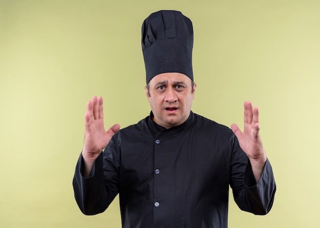 Шеф-повар-мужчина в черной униформе и поварской шляпе, жестикулирующий руками, показывающими знак большого размера, удивлен, измеряет символ, стоящий на зеленом фоне
