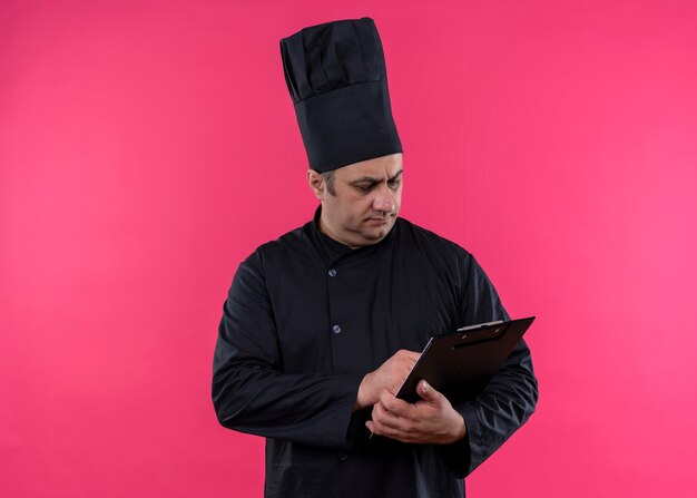 Male chef cook wearing black uniform and cook hat holding clipboard looking at it with serious face standing over pink background