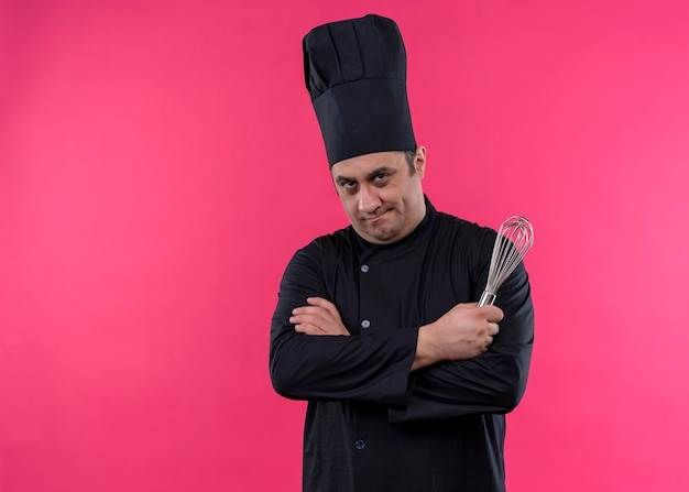 Male chef cook wearing black uniform and cook hat holding corolla looking at camera with serious face with crossed arms standing over pink background