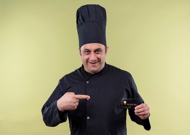 남성 요리사는 검은 색 유니폼을 입고 요리하고 녹색 배경 위에 유쾌하게 서있는 미소를 손가락으로 가리키는 신용 카드를 들고 모자를 요리합니다.