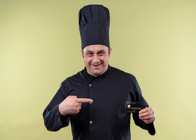 黒のユニフォームを着て、緑の背景の上に元気に立って笑ってそれに指で指しているクレジットカードを保持している帽子を調理する男性シェフ