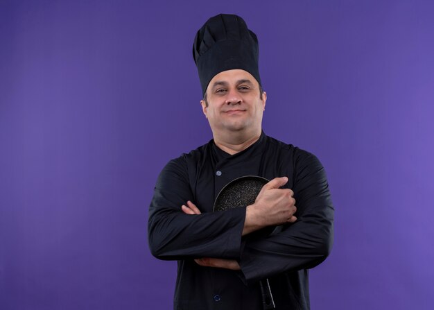 남성 요리사는 검은 색 유니폼을 입고 요리사 모자를 들고 보라색 배경 위에 서있는 자신감있는 미소로 카메라를보고 프라이팬을 요리합니다.