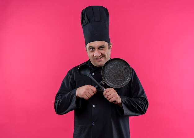 Male chef cook wearing black uniform and cook hat holding frying pan and spoon crossing hands looking at camera with smile standing over pink background