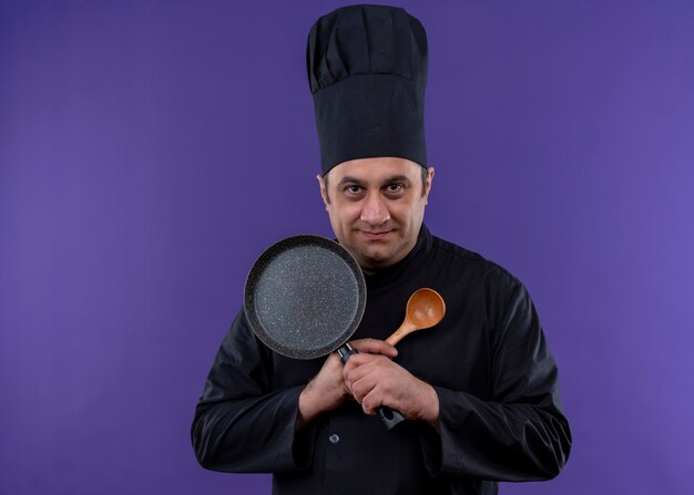 Male chef cook wearing black uniform and cook hat holding frying pan and wooden spoon crossing hands looking at camera smiling standing over purple background