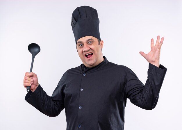 Male chef cook wearing black uniform and cook hat holding ladle raising arm looking at camera with aggressive expression standing over white background