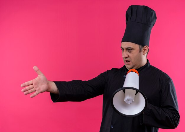 Шеф-повар-мужчина в черной униформе и поварской шляпе держит мегафон с вытянутой рукой и задает вопрос, стоя на розовом фоне