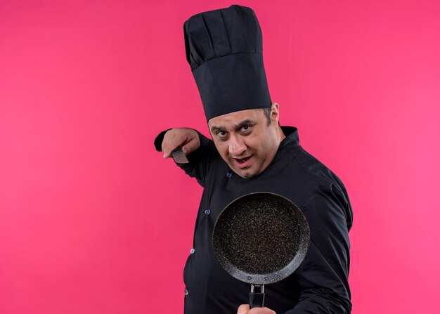 Шеф-повар-мужчина в черной униформе и поварской шляпе держит кастрюлю и нож, глядя в камеру с серьезным лицом, стоящим на розовом фоне