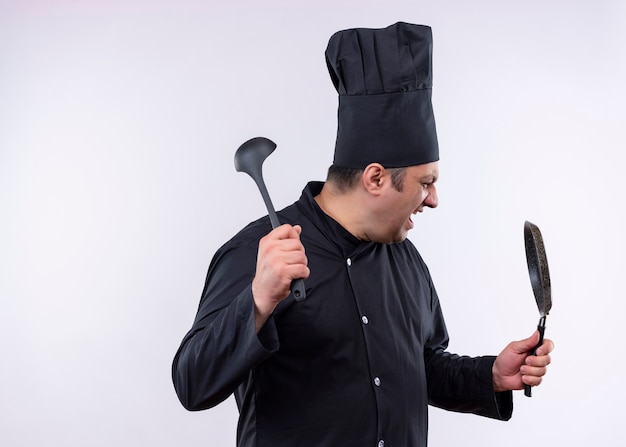 Male chef cook wearing black uniform and cook hat holding pan and ladle shouting with very angry face standing over white background