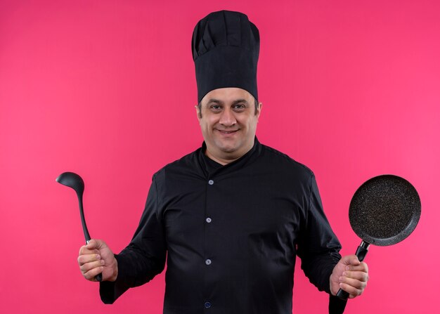 Male chef cook wearing black uniform and cook hat holding pan and ladle smiling confident standing over pink background