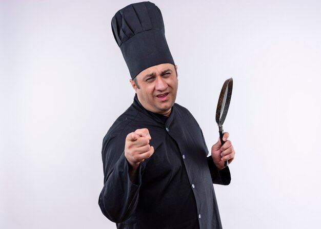 Male chef cook wearing black uniform and cook hat holding a pan pointing with finger to camera happy and positive standing over white background