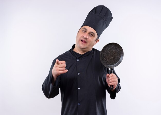 Шеф-повар-мужчина в черной униформе и поварской шляпе держит сковороду, указывая пальцем на камеру, счастливым и позитивным положением на белом фоне