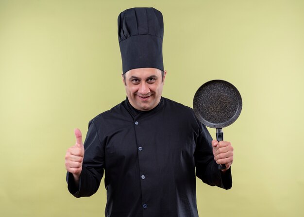 Male chef cook wearing black uniform and cook hat holding a pan showing thumbs up smiling standing over green background