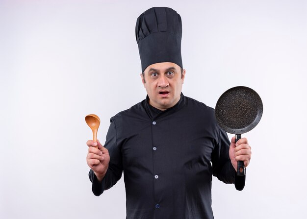 黒の制服を着て、白い背景の上に立って驚いたカメラを見て鍋と木のスプーンを保持している帽子を調理する男性シェフ