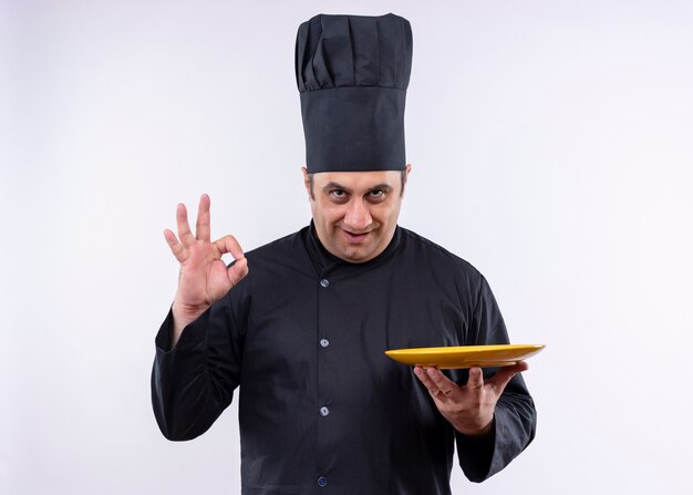 남성 요리사는 검은 색 유니폼을 입고 요리하고 흰색 배경 위에 서있는 확인 서명을 보여주는 접시를 들고 모자를 요리