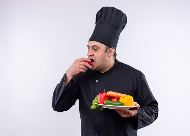 Male chef cook wearing black uniform and cook hat holding plate with fresh vegetables biting tomato standing over white background