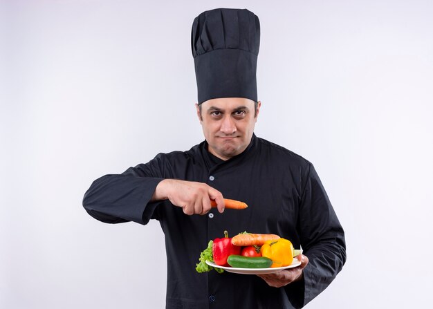 Male chef cook wearing black uniform and cook hat holding plate with fresh vegetables and carrot looking at camera with serious face standing over white background