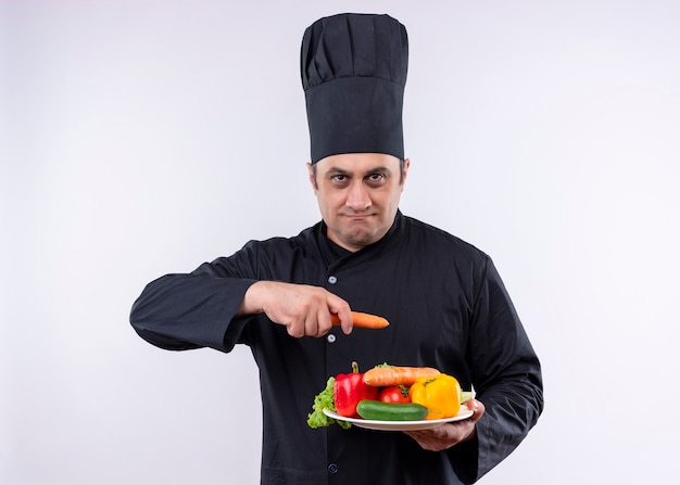 Шеф-повар-мужчина в черной форме и поварской шляпе держит тарелку со свежими овощами и морковью, глядя в камеру с серьезным лицом, стоящим на белом фоне