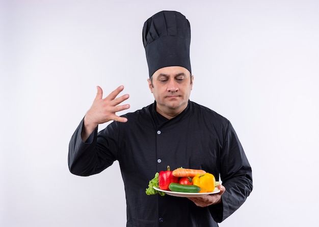Шеф-повар-мужчина в черной униформе и поварской шляпе держит тарелку со свежими овощами, вдыхая аромат свежих овощей, стоящих на белом фоне