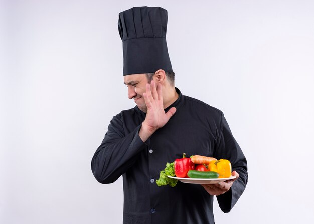 男性シェフの料理人は黒い制服を着て、白い背景の上に立っている嫌な表情で脇を見て新鮮な野菜とプレートを保持している帽子を調理します