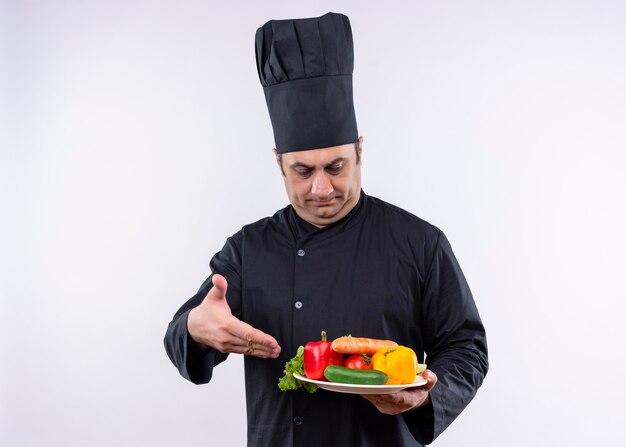 Male chef cook wearing black uniform and cook hat holding plate with fresh vegetables presenting with arm of his hand standing over white background