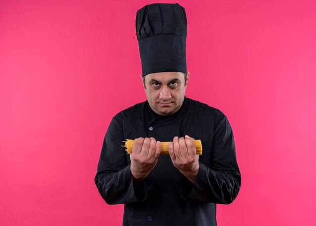 Шеф-повар-мужчина в черной униформе и поварской шляпе держит сырые спагетти, глядя в камеру с серьезным лицом, стоящим на розовом фоне