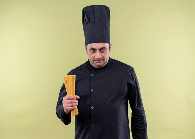 Male chef cook wearing black uniform and cook hat holding row spaghetti looking at camera with serious face standing over green background