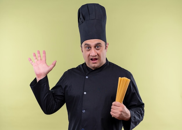 Male chef cook wearing black uniform and cook hat holding row spaghetti looking surprised and amazed with raised hand standing over green background