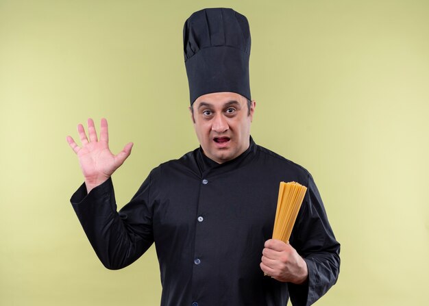 남성 요리사는 검은 색 유니폼을 입고 요리사 모자를 들고 행 스파게티를 들고 놀라게하고 녹색 배경 위에 서있는 손으로 놀랐습니다.