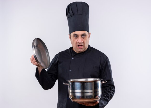 Male chef cook wearing black uniform and cook hat holding saucepan looking at camera cofused standing over white background