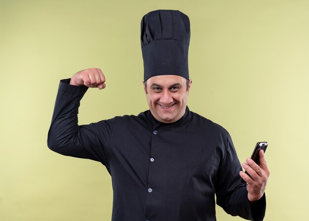 남성 요리사는 검은 색 유니폼을 입고 요리사 모자를 들고 스마트 폰 떨림 주먹을 들고 흥분하고 행복하게 서 녹색 배경 위에 서